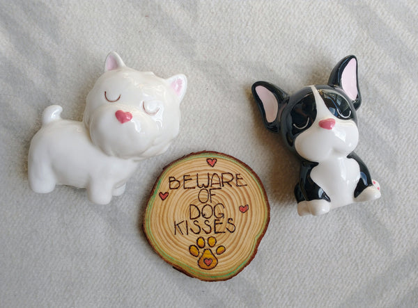 A wooden ornament that has Beware of Dog Kisses wood burned onto it, it is also painted, and is lying on a white background. It is surrounded by two small ceramic dog figurines.