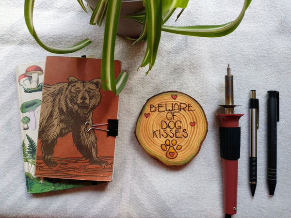 A staged scene with a wooden ornament in the center. It has Beware of Dog Kisses wood burned into it, and it is painted. It is surrounded by a green plant, two small notebooks on the left, and wood burning insturments on the right.