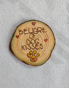 A wood slice coaster that has Beware of Dog Kisses wood burned onto it.