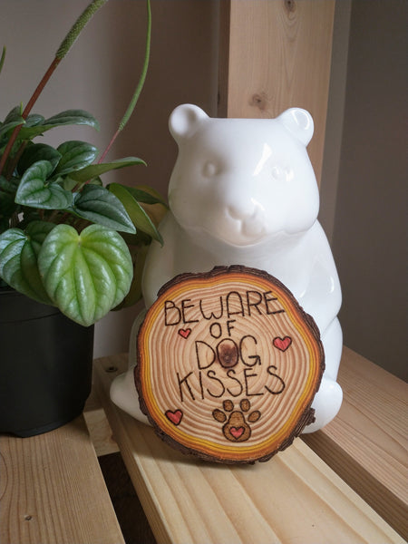 A Beware of Dog Kisses Custom Ornament staged on a wooden shelf. It is leaning against a white ceramic bear figurine, and there is a green plant on the left.