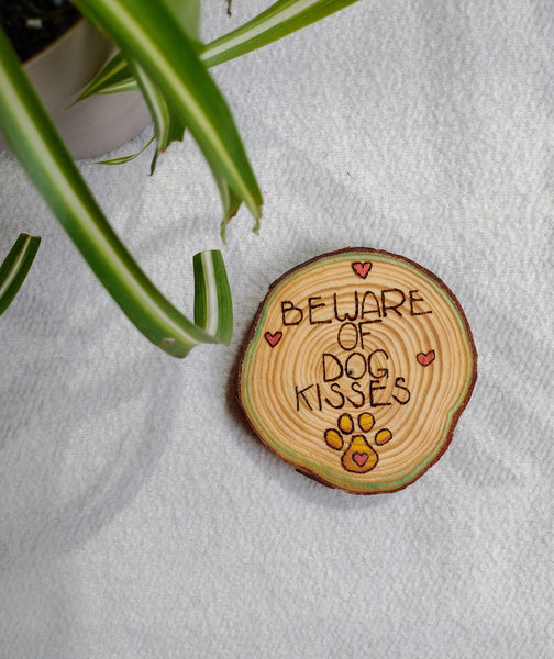 A Beware of Dog Kisses Custom Ornament, which is a design wood burned onto a slice of wood, on a white background with a green plant in the corner