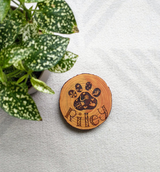 A Custom Cat Paw Print Wood Slice Ornament on a white background, with a green plant in one corner.