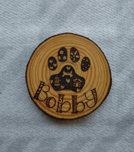 A coaster made of a slice of wood that has a dog paw and the name Bobby wood burned into it.