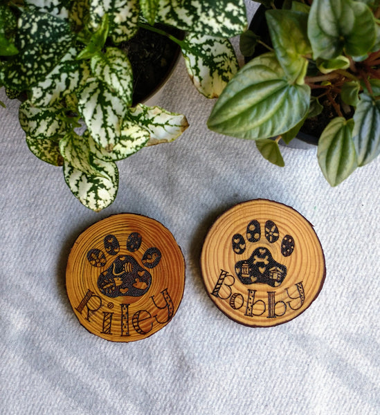 A Custom Cat Paw Print Wood Slice Ornament next to a Custom Dog Paw Print Wood Slice Ornament. They are on a white background with two green plants at the top.