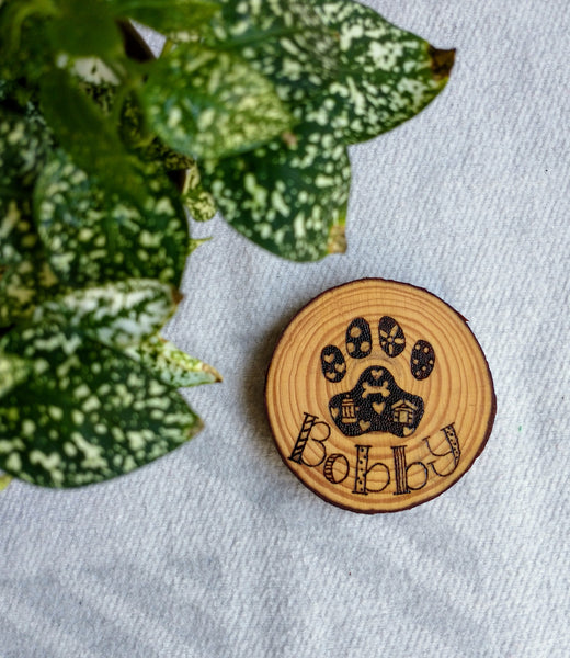 A beautifully wood burned Custom Dog Paw Print Wood Slice Ornament on a white background with a green plant in the corner.