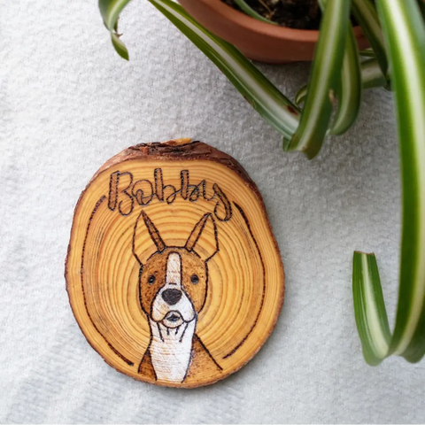 A Custom Pet Portrait Wood Slice Ornament that has a boston terrier and the name Bobby wood burned onto it. It is on a white background with a green plant in the corner.