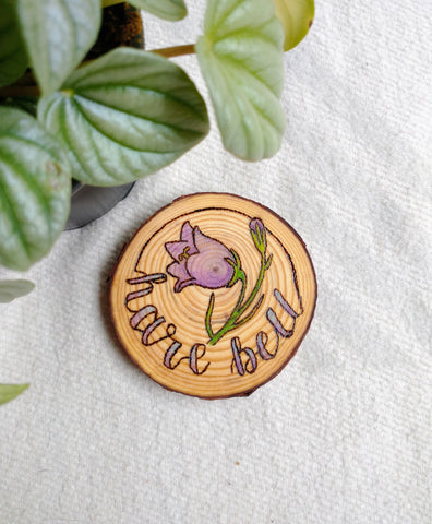 A Harebell Unique Woodburned Wood Slice Ornament on a white background with a green plant in the corner. The Harebell design is woodburned into the ornament, with purple and green paint added for color.