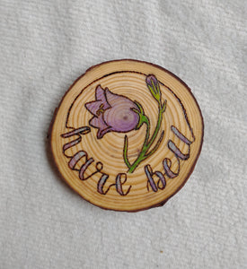 A wood slice coaster with a Harebell Alberta Wildflower design wood-burned into it. There is purple and green paint added for color.