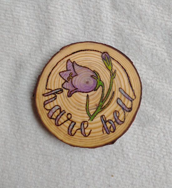 A wood slice coaster with a Harebell Alberta Wildflower design wood-burned into it. There is purple and green paint added for color.