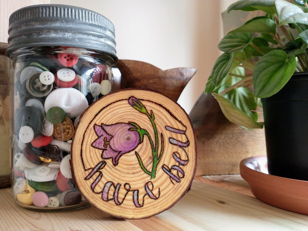 A Harebell Ornament staged on a wooden shelf next to a green plant and an old jar of mismatched buttons.