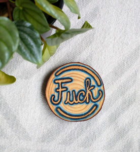 A Unique Fuck Wood-burned Wood Slice Ornament lying on a white background with a green plant in the corner.