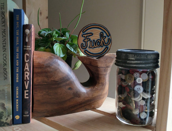A Fuck Wood-burned Wood Slice Ornament staged on a wooden shelf. It is balancing on a bookend, surrounded by books, a green plant, and a jar of mismatched buttons.