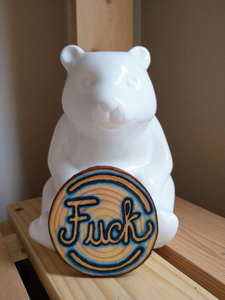 A Fuck Ornament in front of a white ceramic figurine of a sitting bear. 